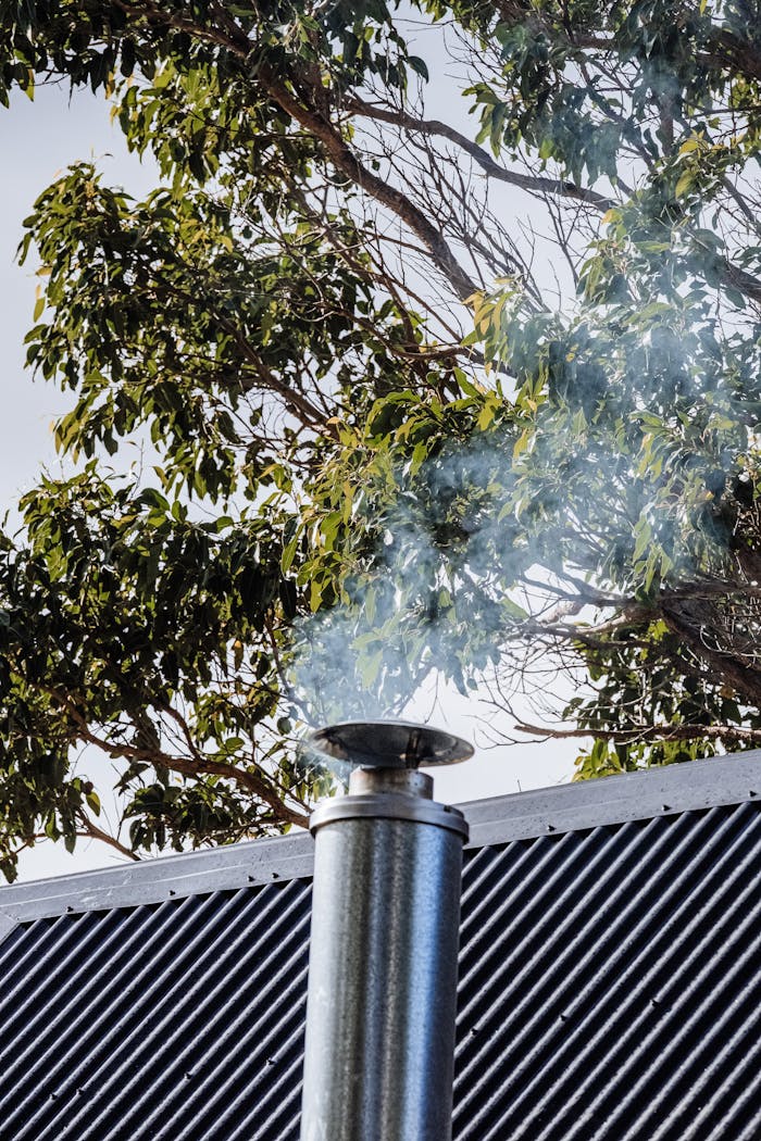 about-02 Vertical shot of a chimney releasing smoke with leafy trees in the background, ideal for environmental themes.